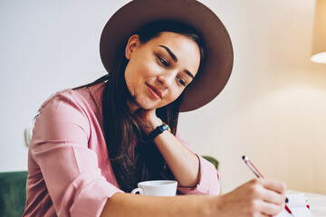 Attractive young woman in trendy hat writing in diary resting in cozy coffee shop interior,beautiful female student noting creative ideas and drawing graphic in sketchbook for project planning
