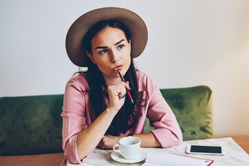 Skilled female designer pondering on crating startup strategy concentrated on ideas sitting with pen and notepad in cafe, cute hipster girl in stylish apparel thinking while making to do list