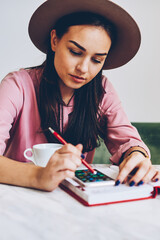 Pensive young woman in trendy hat installing application on smartphone for organizing day sitting in cafe,female student texting message on mobile phone chatting in social network during break