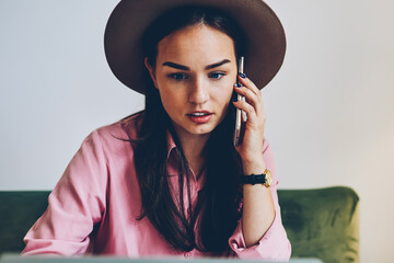 Pensive female freelancer checking banking account online and talking with customer support operator on telephone, young woman having mobile conversation reading news about her business on laptop