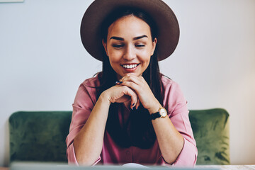 Smiling young woman in stylish hat watching online movie using laptop computer during free time, positive female satisfied with freelance job reading news on netbook spending time in cafe interior