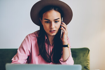 Pensive female freelancer checking banking account online and talking with customer support operator on telephone, young woman having mobile conversation reading news about her business on laptop