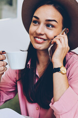 Attractive brunette hipster girl resting in cafe interior talking on mobile phone with friend, smiling young woman in trendy hat having telephone conversation enjoying cup of favorite coffee