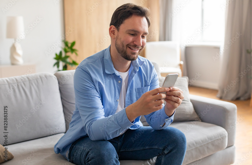 Poster A bearded man is seated on a couch, engrossed in his smartphone screen. He appears focused as he scrolls and interacts with the device, surrounded by a living room setting.