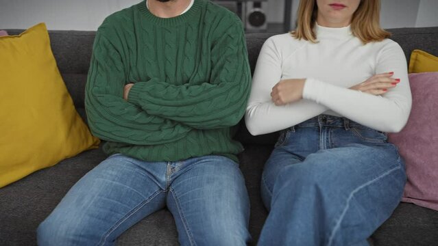 A man and woman sit apart on a sofa, embodying tension in an indoor domestic setting, with crossed arms and neutral expressions against colorful cushions.
