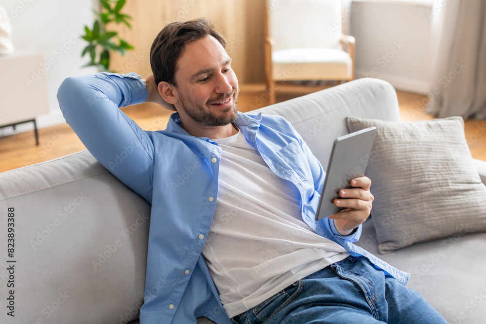 Poster A man is casually sitting on a couch, engrossed in using a tablet device. He appears focused as he interacts with the screen, showcasing modern technology in everyday life.