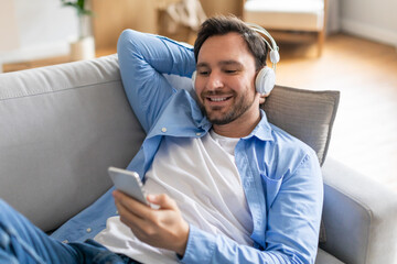 A man is sitting on a couch while listening to music on his phone. He appears relaxed and engaged in the music playing through his headphones.