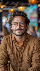Obraz premium A young man with glasses and a brown shirt smiles warmly while sitting in a vibrant caf? with colorful lights and a lively atmosphere in the background
