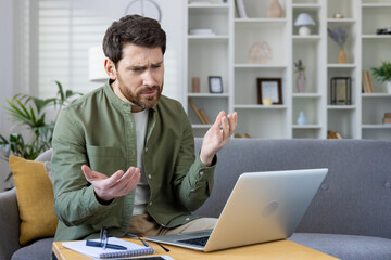 Frustrated man working from home on laptop with confused expression. Young professional experiencing technical difficulties while working remotely in a modern living room setup.