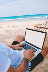 Pensive man freelancer in sunglasses texting on keyboard of modern laptop working remotely during summer vacations on sea shore. Male tourist businessman resting on hammock at distance job on computer
