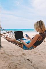 Young woman tourist reading notification on smartphone in roaming while lying on hammock and working remote at laptop computer with mock up area for your content during summer vacation on ocean beach