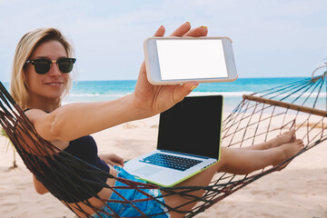 Cheerful blonde female freelancer showing smartphone with blank screen for your internet web page while working at netbook on hammock during vacations distance job on tropical sea shore in summer time