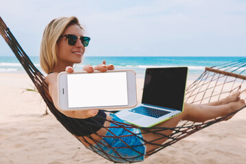 Female tourist freelancer showing smartphone with blank screen for application or entrepreneur job website while working remotely at modern laptop computer during summer vacation holidays on beach