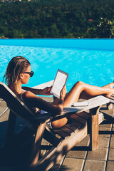 Sexy young woman sunbathing on sunbed near blue pool while reading news on websites using good 4G...