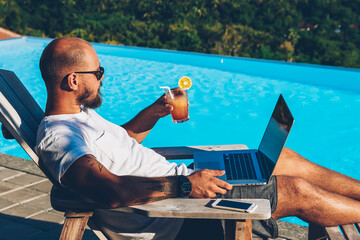 Bearded businessman in sunglasses enjoying cocktail while watching videos online on websites on...