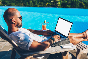 Businessman enjoying hot tour during summer resort lying on sunbed with cocktail near pool while earning money doing distance job at netbook with blank copy space screen for your advertising content