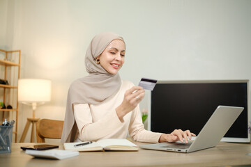 A woman wearing a hijab is sitting at a desk with a laptop and a credit card. She is smiling as she...