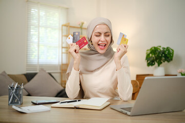 A woman is holding up a stack of credit cards and smiling. She is sitting at a desk with a laptop and a notebook