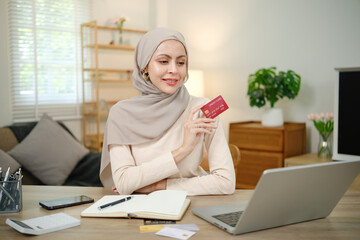 A woman wearing a head scarf is holding a red Visa card. She is sitting at a desk with a laptop and a notebook