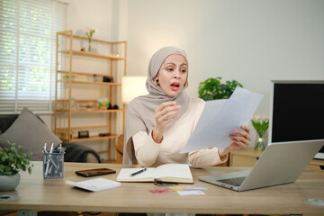 A woman wearing a hijab is sitting at a desk with a laptop and a piece of paper. She is looking at the paper and she is surprised