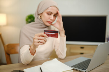 A woman is holding a credit card and looking at a laptop. She is in a state of distress or worry