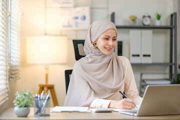 A woman wearing a head scarf is sitting at a desk with a laptop and a notebook. She is smiling and...