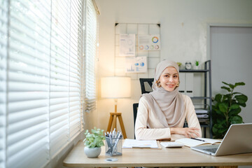A woman wearing a head scarf sits at a desk with a laptop and a stack of papers. She is focused on her work, possibly preparing for a presentation or meeting. The room is well-lit