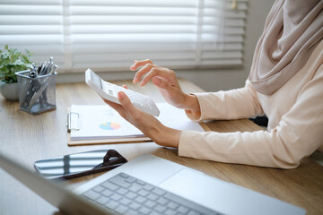 A woman is sitting at a desk with a calculator and a laptop. She is focused on her work and she is...