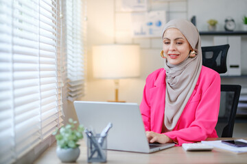 A woman wearing a pink jacket and a head scarf is sitting at a desk with a laptop. She is smiling and she is enjoying her work