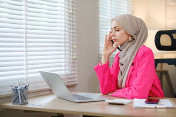 A woman in a pink jacket is talking on her cell phone while sitting at a desk. She is wearing a head scarf and she is in a professional setting