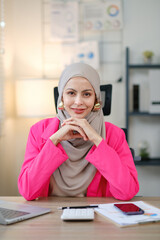 A woman wearing a pink jacket and a grey scarf sits at a desk with a laptop, a cell phone, and a calculator. She is smiling and she is in a positive mood