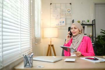 A woman in a pink jacket is talking on her cell phone while sitting at a desk. She is wearing a hijab and she is in a professional setting
