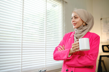 A woman in a pink jacket is holding a white coffee cup. She is smiling and looking out the window