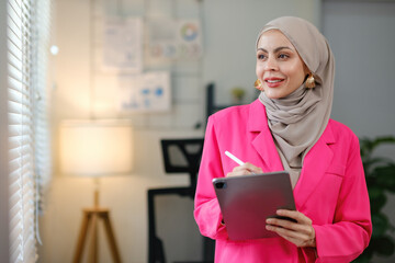 A woman in a pink jacket is holding a tablet and writing on a clipboard. She is focused and determined