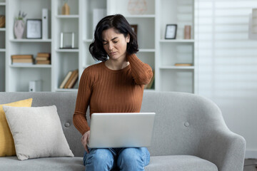 Photo of a young woman sitting at home on the sofa, holding a laptop on her lap, working and...