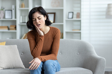 Obraz premium A sick young woman sits at home on the sofa and holds her hand to her cheek, grimaced from severe toothache, needs medical help from a dentist