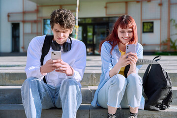Teenage youth guy and girl college students sitting outdoor using smartphone