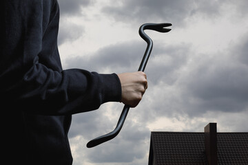 A thief with a crowbar in front of a sky and the roof of a house.