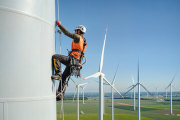 A worker in safety gear climbing a wind turbine with a field of wind turbines in the background.