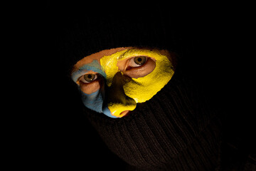 The face of a girl in a black balaclava and the flag of Ukraine is painted on her face with paints