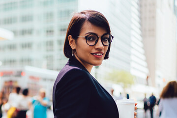 Half length portrait of happy businesswoman dressed in stylish formal wear and eyeglasses smiling at camera holding coffee to go standing on office building background near publicity area