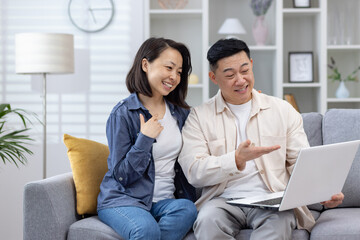 Happy asian couple smiling and enjoying a video call on their laptop while sitting on a sofa at home.