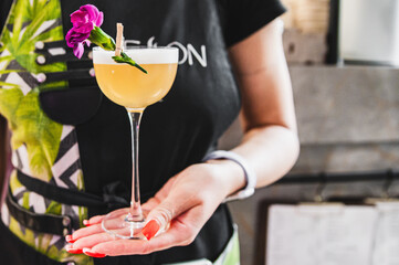 A person’s torso holds a stemmed glass with an orange beverage, garnished with a pink flower and...