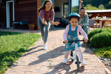 Small girl riding bicycle with support of her parents outdoors.