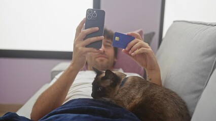 A young man lounges with his cat while shopping online using a smartphone and credit card in a cozy living room.