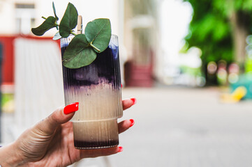 A hand holds a layered beverage in a clear glass, garnished with a green leaf, against a blurred street background.