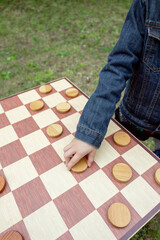 Close-up of a child's hand moving pieces on a wooden checkers board outdoors. Playful and...