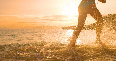 LENS FLARE, CLOSE UP, SILHOUETTE: young woman splashes water while running through shallow seawater...