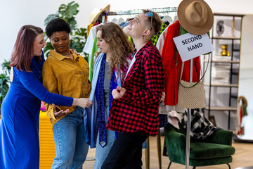 Group of friends generation z girls and boy enjoying shopping in a second hand store, trying hats, jackets, buying vintage brand clothes for low prices with discounts. Flea market, bargain hunting