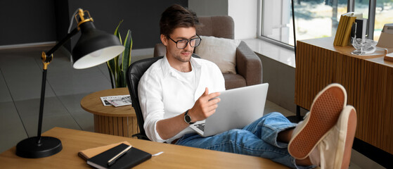 Handsome young man with wristwatch  and eyeglasses working on laptop having put his legs on table in office